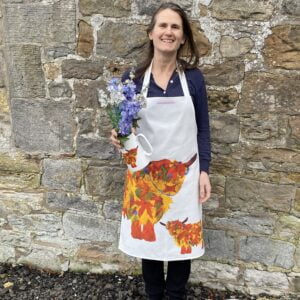 Highland Cow Apron with a large bold highland cow and baby print in autumnal orange russet colours. Worn by Chloe holding a jug of freshly picked flowers
