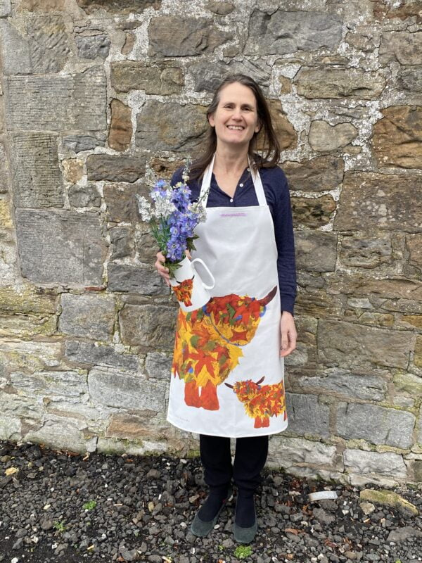Highland Cow Apron with a large bold highland cow and baby print in autumnal orange russet colours. Worn by Chloe holding a jug of freshly picked flowers